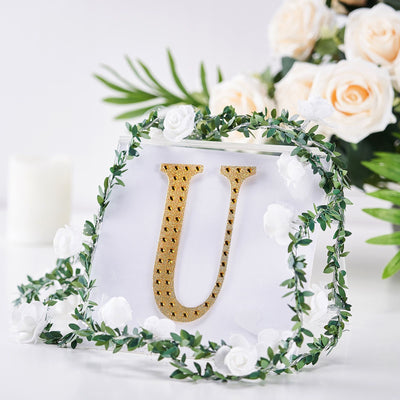 Gold letter "U" surrounded by faux flowers and greenery on a white background.