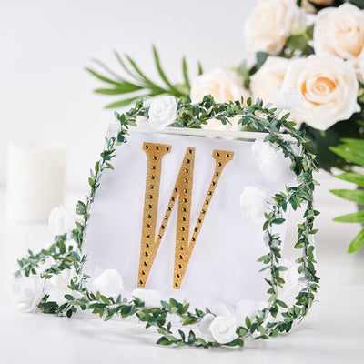 Gold letter "W" decoration surrounded by greenery and white roses.