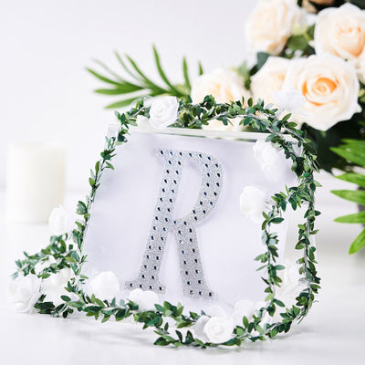 Decorative letter "R" with rhinestones surrounded by greenery and flowers.