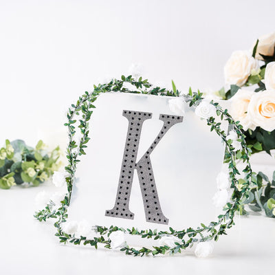 Decorative letter "K" surrounded by a floral wreath on a plain background.