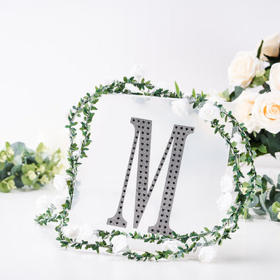 Decorative letter "M" embellished with rhinestones, framed by floral greenery.