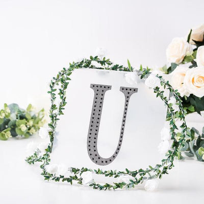Decorative letter "U" with rhinestones surrounded by floral greenery.