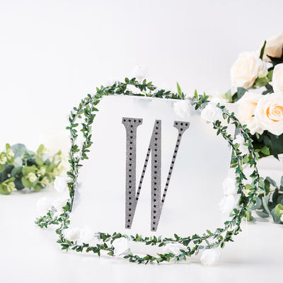 Silver letter "W" surrounded by floral greenery on a clear background.