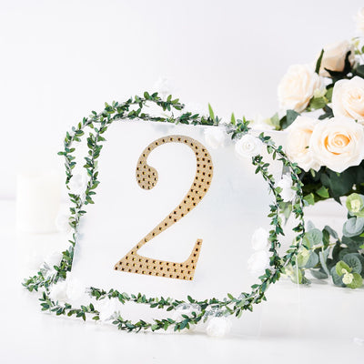Gold number 2 sign adorned with greenery and white flowers on a white backdrop.