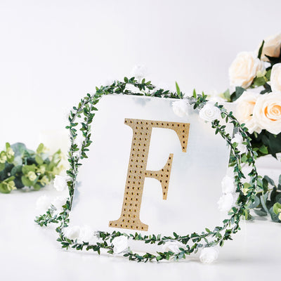 Gold letter F surrounded by greenery and flowers on a white background.