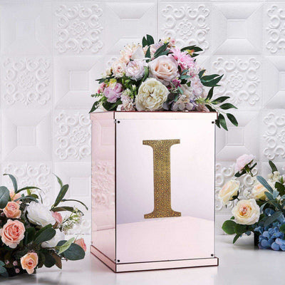Decorative mirrored box with floral arrangement and gold letter "I" on a textured backdrop.