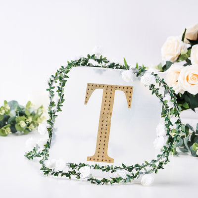 Gold letter "T" adorned with greenery and flowers on a clear background.