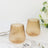 Set of two ribbed amber glass tumblers on a marble table with utensils and flowers.