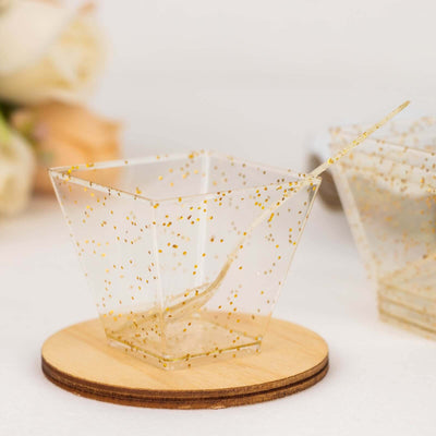 Clear plastic dessert cup with gold glitter dots and a spoon on a wooden coaster.