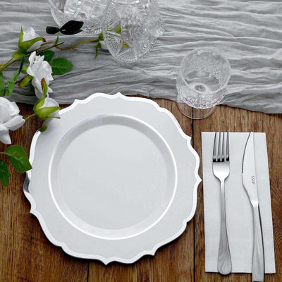 Elegant white plate with scalloped edge, silver cutlery, and glassware on textured fabric.