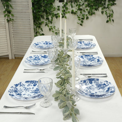 Elegant blue and white dinnerware set arranged on a table with greenery and candles.