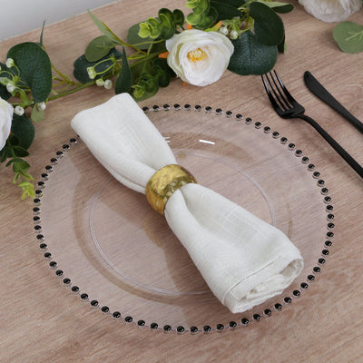 Elegant white napkin with gold ring on a clear plate, surrounded by greenery and flowers.
