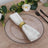 Elegant white napkin with gold ring on a clear plate, surrounded by greenery and flowers.