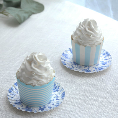 Two decorated cupcakes with whipped cream in striped cups on floral plates.