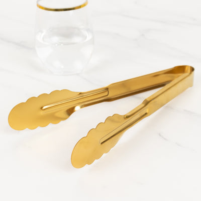Gold kitchen tongs on marble countertop with a glass of water in the background.