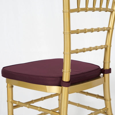 Gold chiavari chair with burgundy cushion on a plain backdrop.