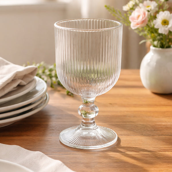 Clear ribbed glass goblet on a wooden table with flowers in the background.