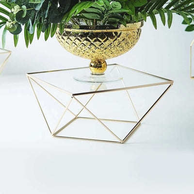 Gold geometric plant stand with glass top and greenery.