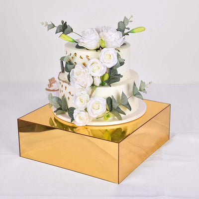 Elegant three-tier cake with white flowers and gold accents on a mirrored base.