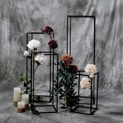 Black metal flower stands with peonies and candles on a textured backdrop.
