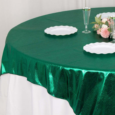 Green shiny tablecloth draping over a round table with white plates and floral centerpiece.