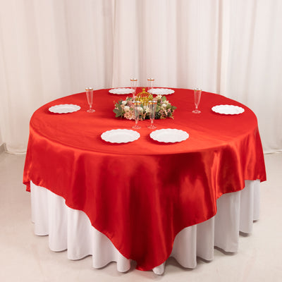 Red satin tablecloth on a round table with plates and floral centerpiece.