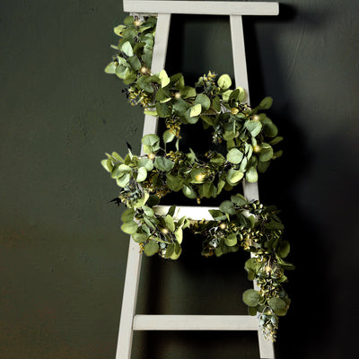 Green eucalyptus wreath hanging on a white ladder against a dark background.