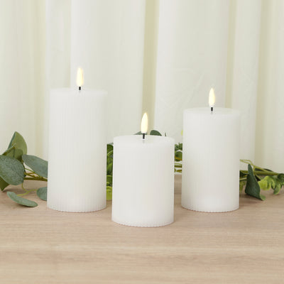 Three white ribbed candles with flames on a wooden surface.
