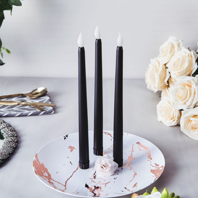 Black tapered candles on a decorative plate with roses in the background.