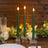 Three lit green taper candles on a rustic wooden table with greenery and glassware