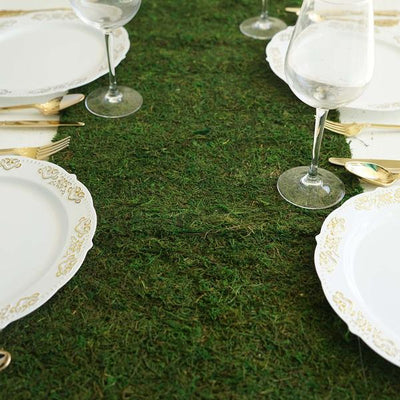 Elegant table setting with gold-accented plates and glassware on a moss backdrop.