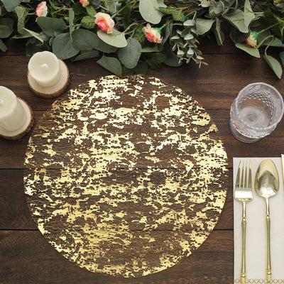 Gold textured round placemat on a rustic wooden table with greenery and dining utensils.