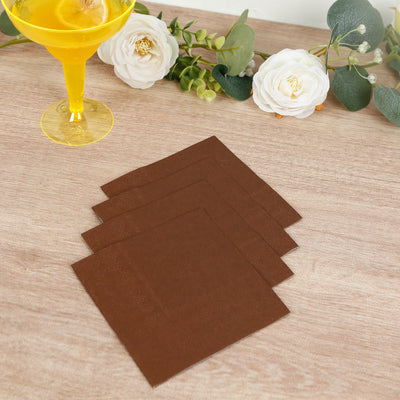 Brown paper napkins stacked on a wooden table with a cocktail and flowers.