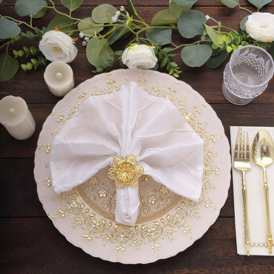 Elegant white napkin with gold flower ring on ornate plate, surrounded by greenery.