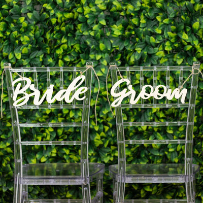 Bride and Groom transparent chairs with decorative signage against a green backdrop.