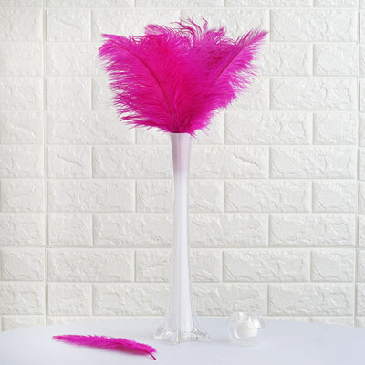 Pink feather arrangement in a tall clear vase against a textured white backdrop.