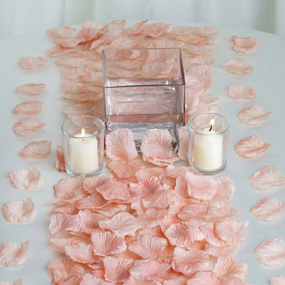 Pink rose petals with candles and a glass vase centerpiece on a white table.