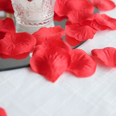 Red silk rose petals scattered around a candle on a table.