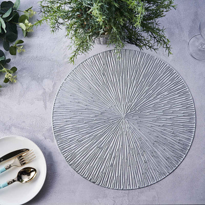 Textured round placemat on a grey surface with greenery and dining utensils.