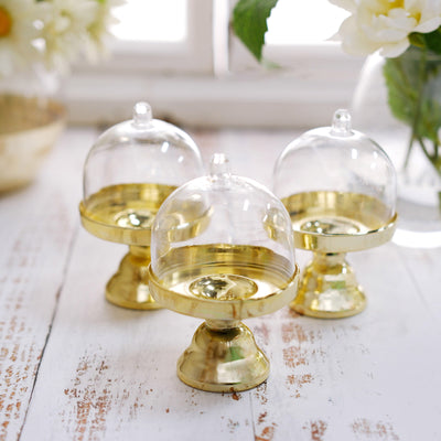 Gold cake stands with glass domes on a rustic wooden surface.