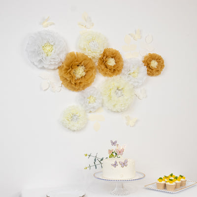 Decorative paper flowers in white, yellow, and gold on a wall with a cake and cupcakes.