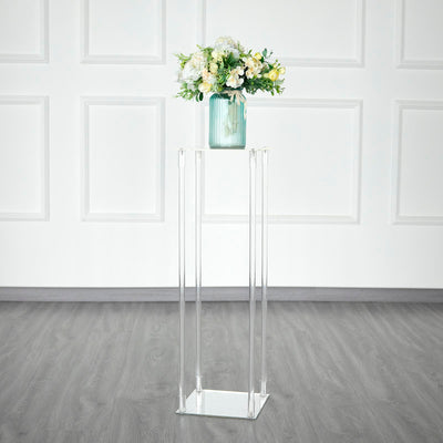 Clear acrylic pedestal with a vase of flowers on a light wood floor.