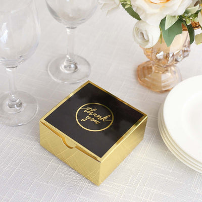 Gold thank you gift box on a table with wine glasses and floral arrangement.