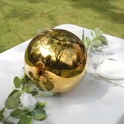 Gold decorative orb on a table with greenery and a glass, reflecting the outdoors.
