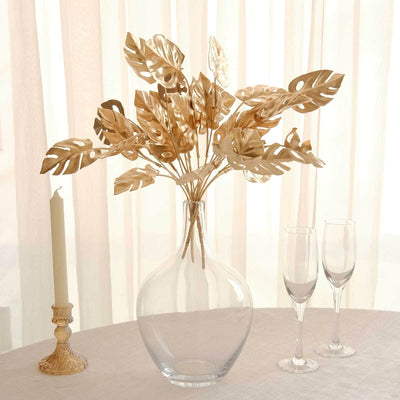 Gold leaf arrangement in a clear vase on a table with candles and glasses.