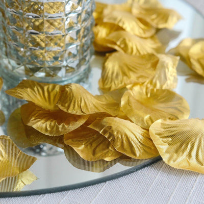 Gold silk rose petal decorations scattered around a glass vase on a reflective surface.