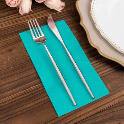 Silver fork and knife on a teal napkin, with a plate and flowers in the background.