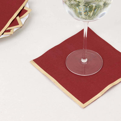 Maroon napkin with gold trim under a glass on a white surface.