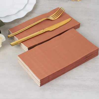 Brown paper napkins stacked with a gold fork and knife on a tabletop.