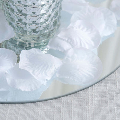 Glass vase surrounded by white silk rose petals on a mirrored surface.